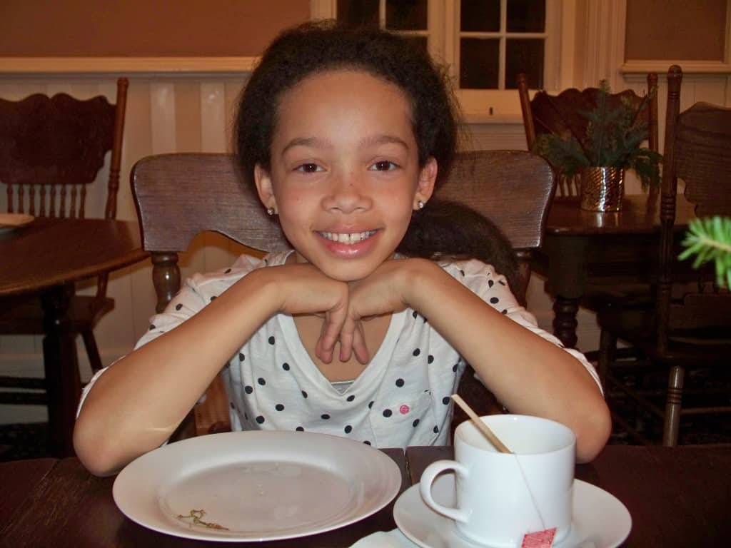 A little girl smiling at the camera while at breakfast. In front of her on the table is an empty plate and a mug of tea